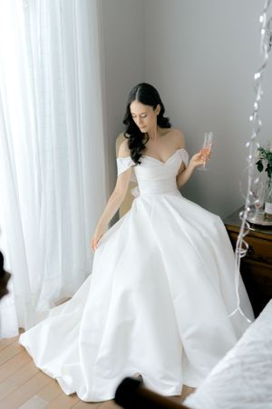 A bride in an elegant white off-shoulder ball gown holds a champagne glass while standing by sheer curtains.