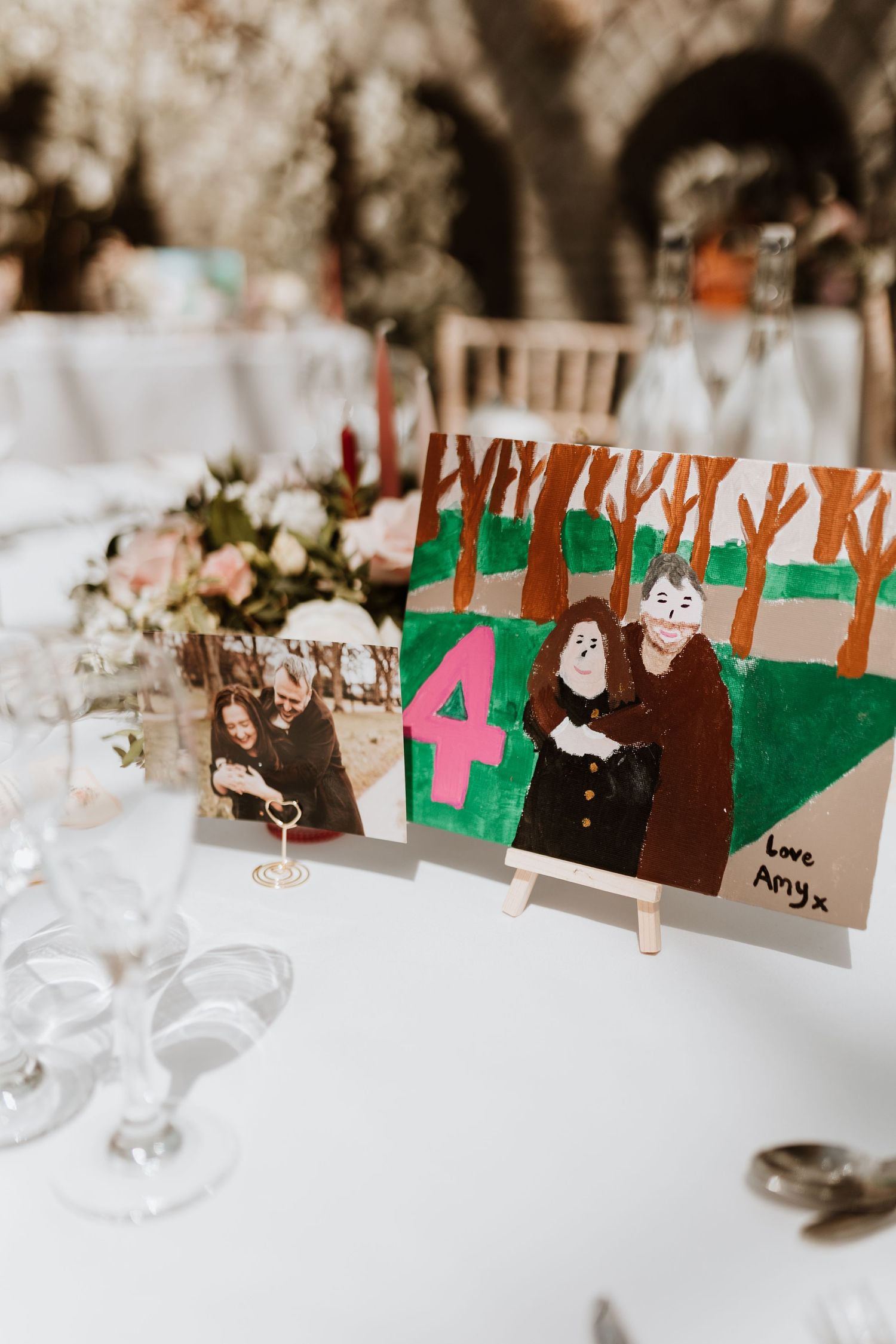 A creative table number display with a painted illustration and photo on a white tablecloth at a wedding reception.