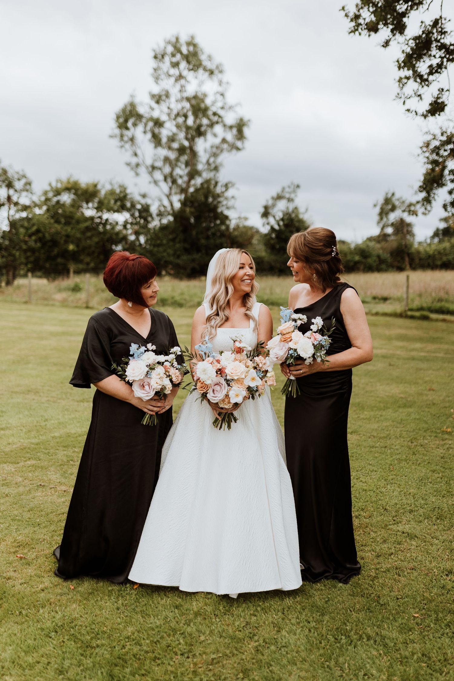 Three women pose together outdoors at a wedding, with the bride in white and bridesmaids in black holding floral bouquets.