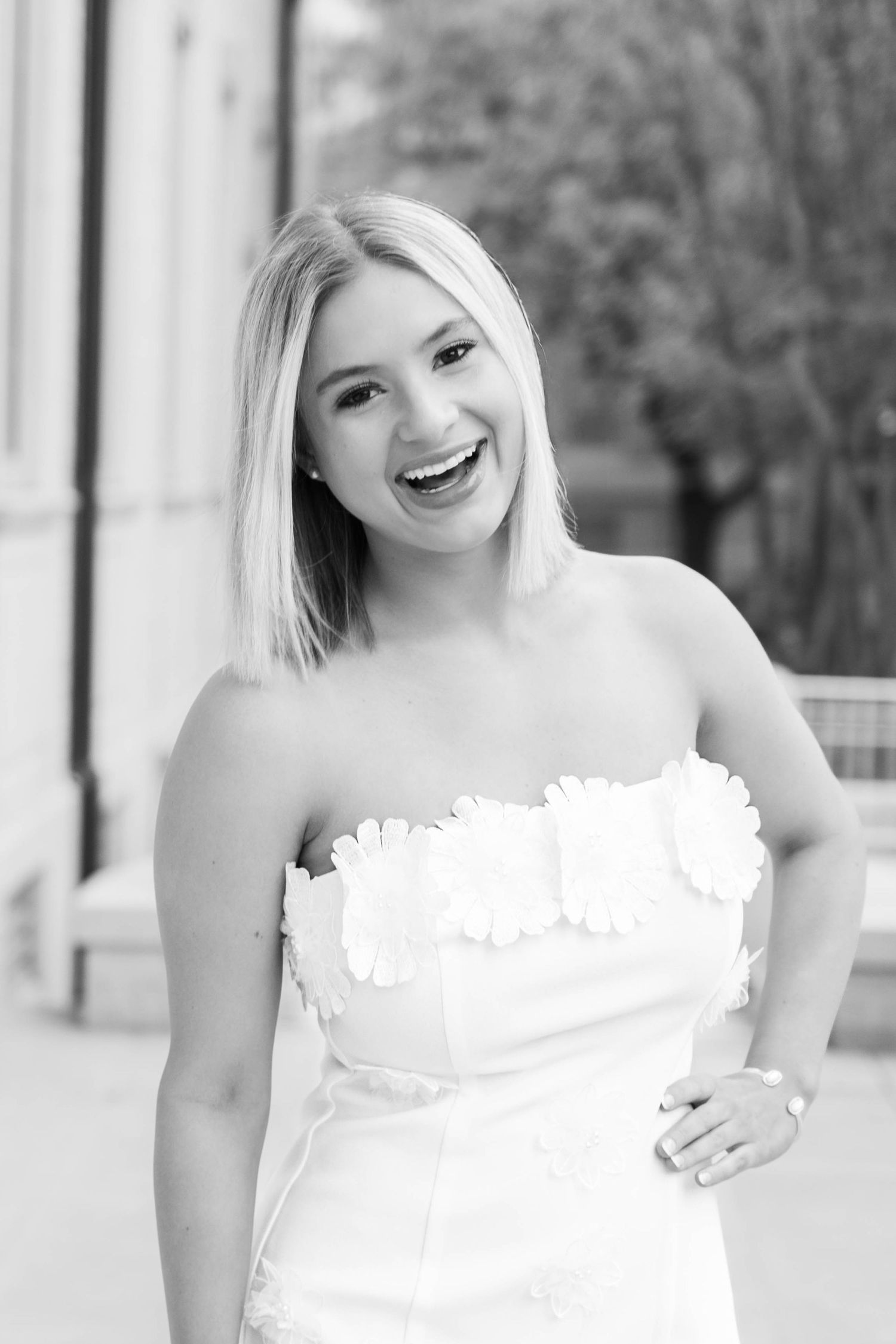 A black and white portrait of a joyful smile while wearing a white strapless dress outdoors.