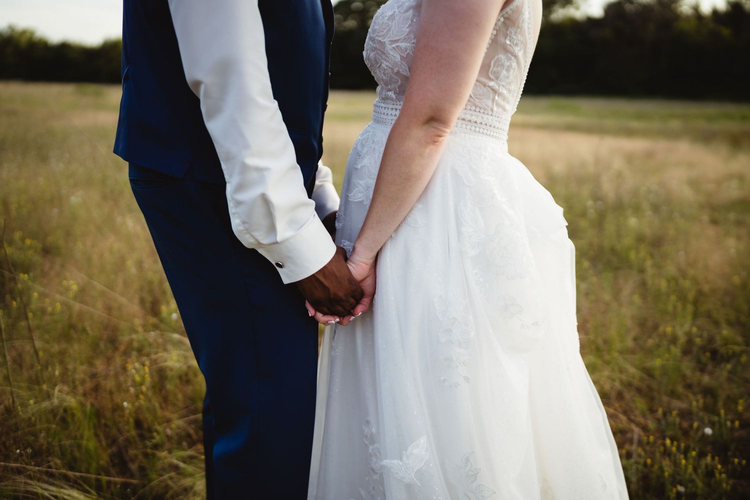 Close-up view of newlyweds holding hands in a field at sunset, bride in white gown and groom in navy suit.