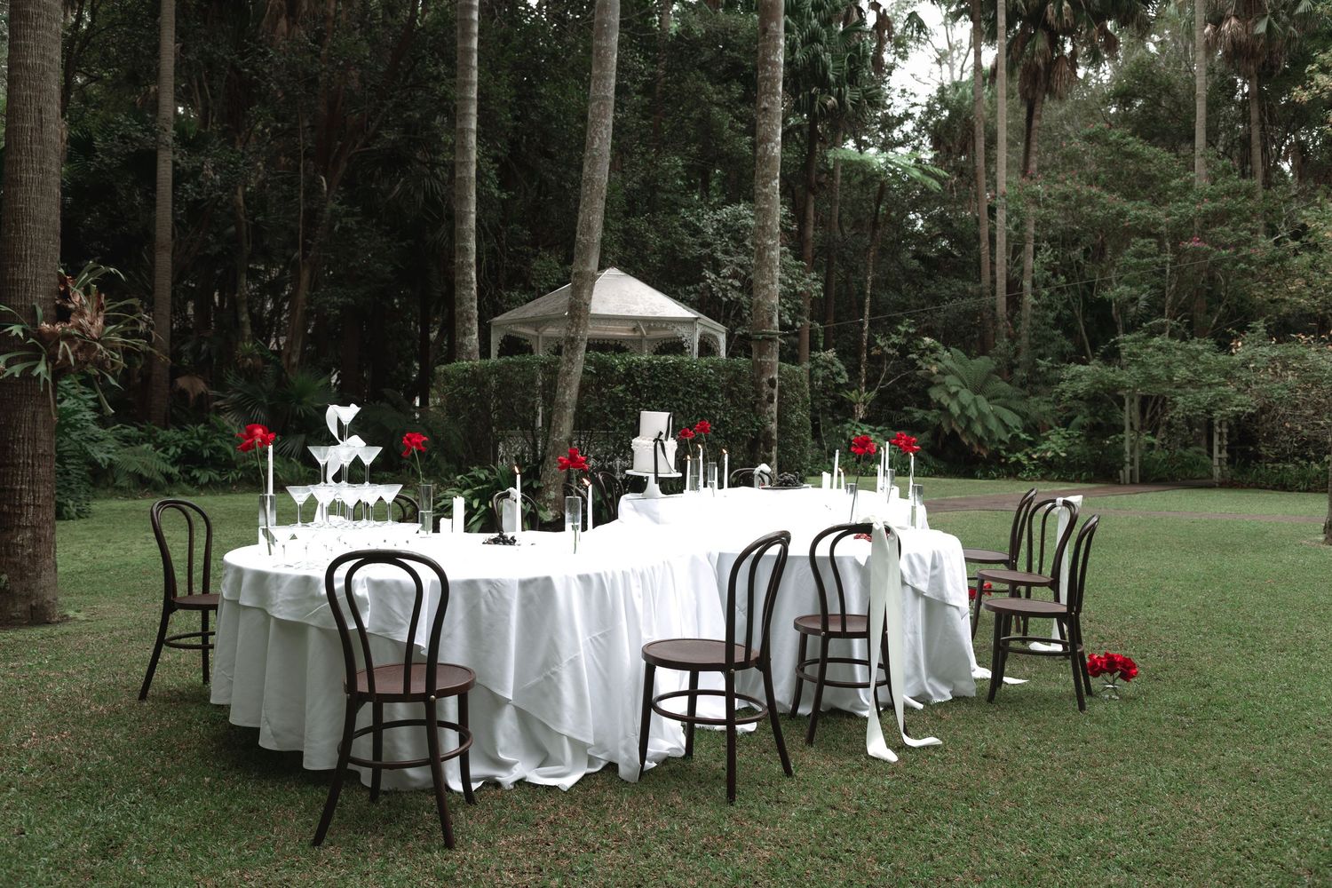 Tables with white tablecloths and chairs set up in a wooded outdoor area for an elegant garden event.