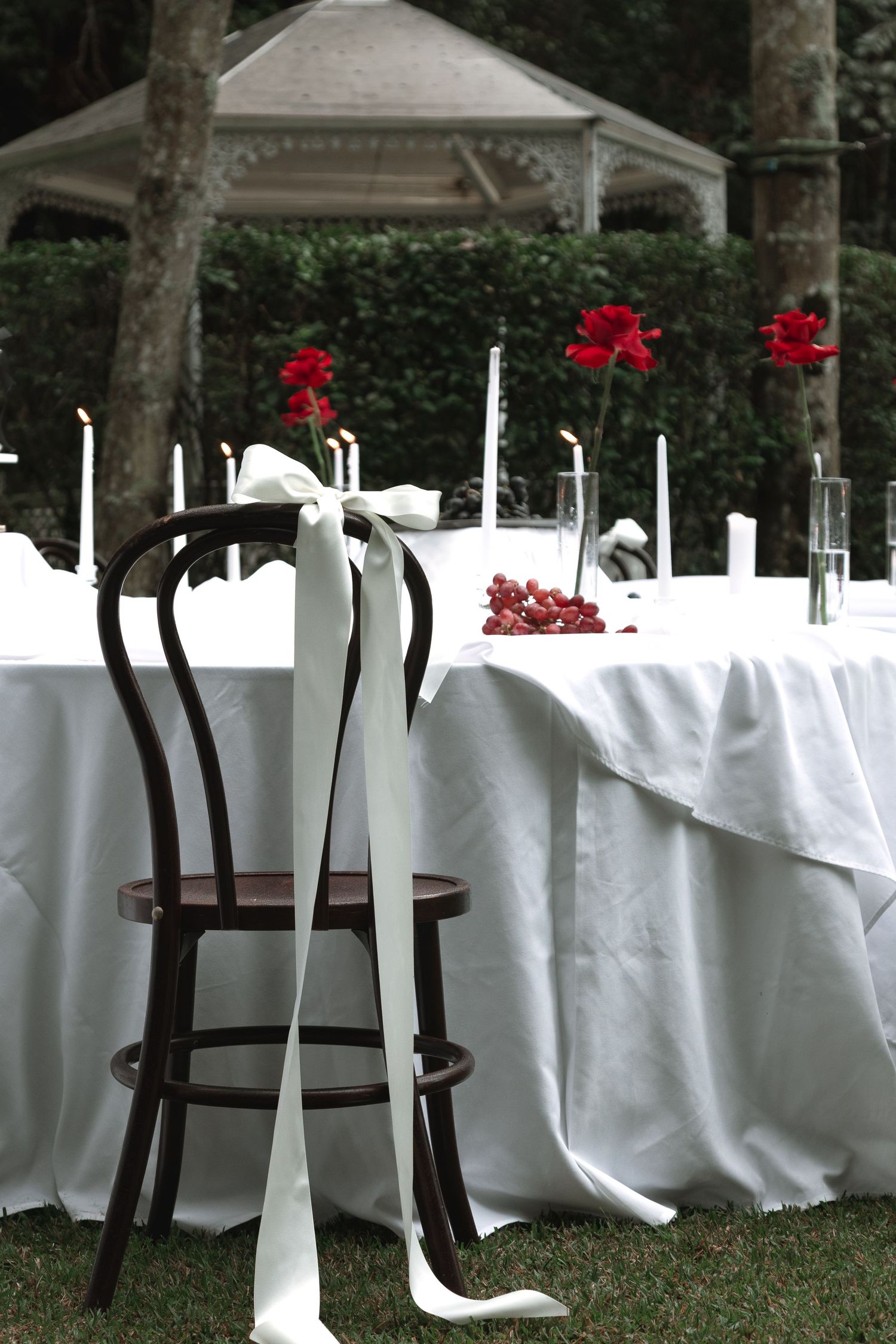 Elegant outdoor wedding table setting with white linens, bentwood chair, and red roses under a gazebo in a garden.
