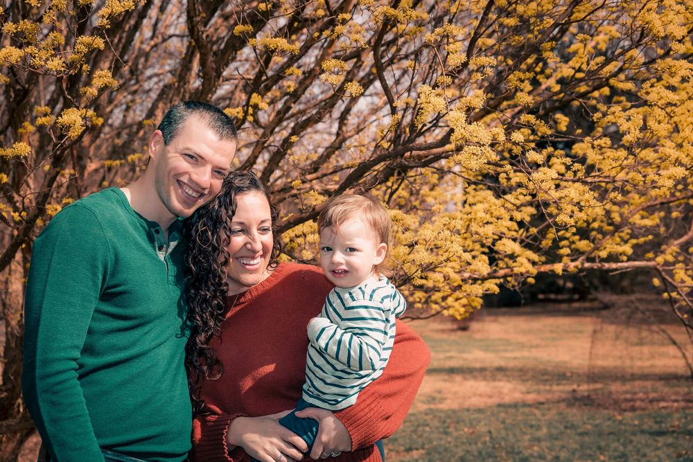 A family stands together outdoors on an autumn day with golden yellow foliage in the background.