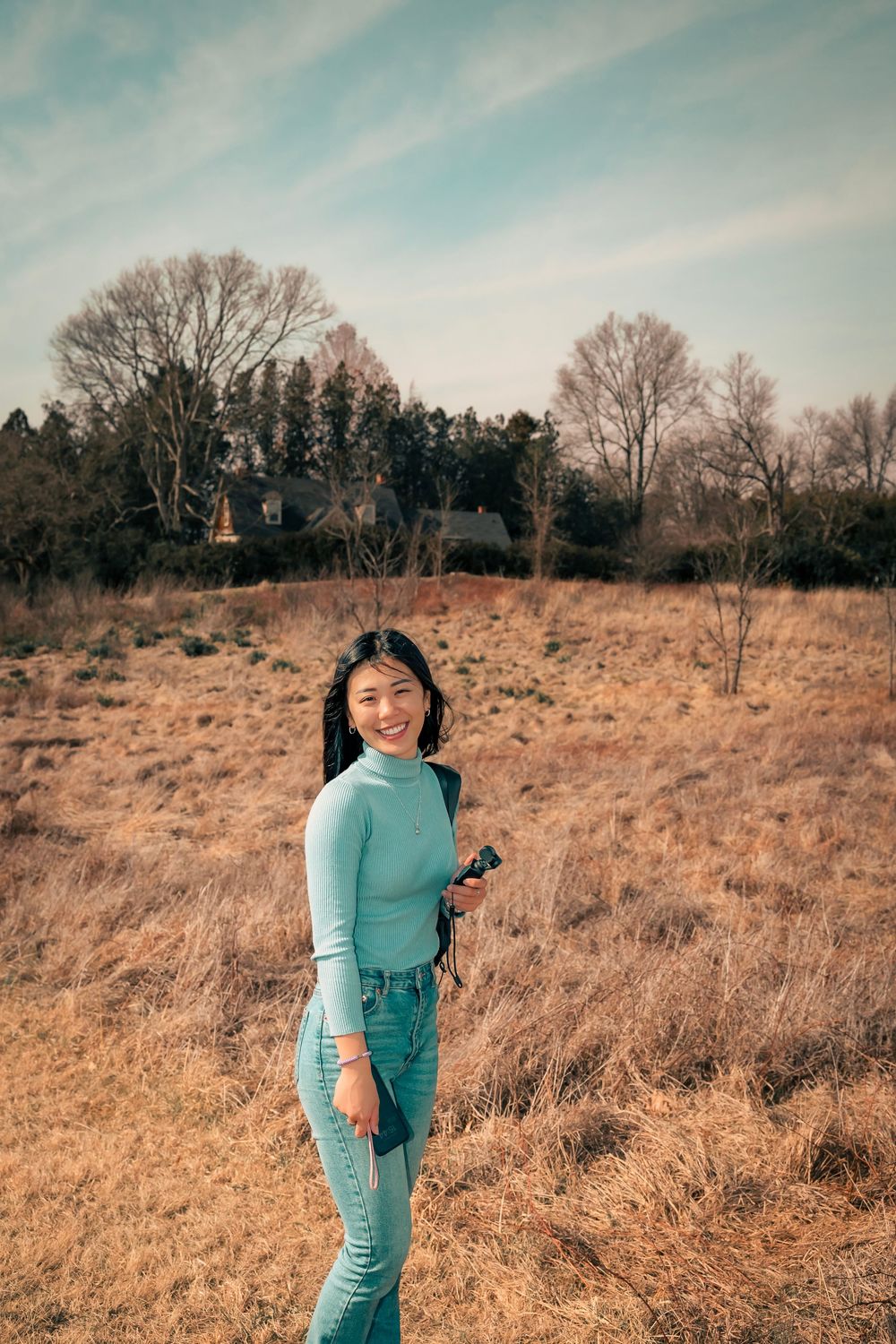 Person wearing turquoise sweater and jeans stands in a golden autumn field with bare trees in the background.