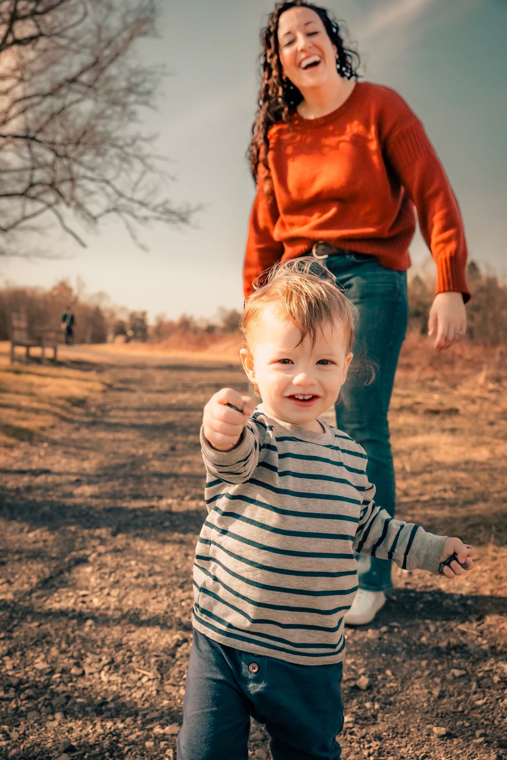 A toddler in a striped shirt plays outdoors on an autumn day while accompanied by someone in an orange sweater.