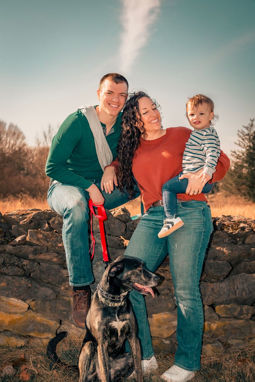 Outdoor family photo session shows couple in casual wear with small child and black dog against stone wall at sunset.