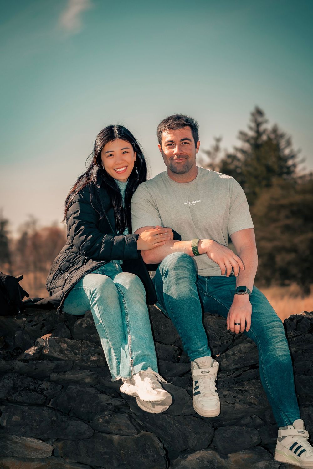 A couple sits together on a rock formation wearing casual outfits and sneakers during sunset.