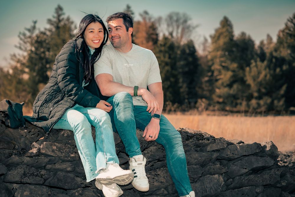A couple in casual wear sits together on dark rocks with evergreen trees in the background during golden hour.