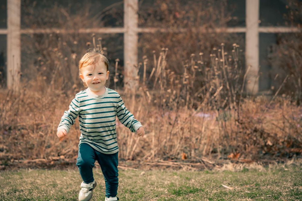 A joyful toddler in a striped shirt runs and plays in an outdoor setting with fall foliage in the background.