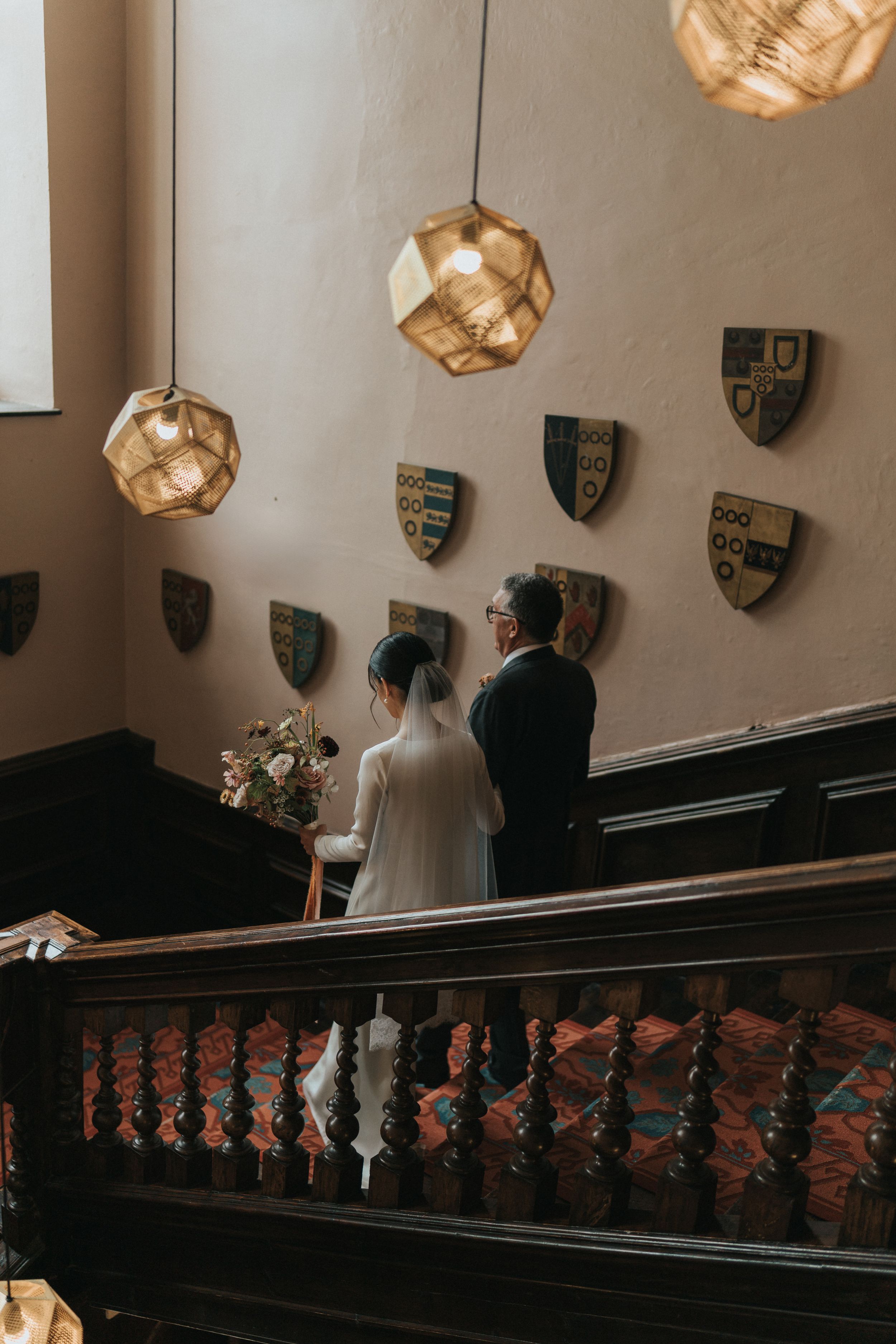 A couple walking up grand staircase with ornate wooden railings and decorative shields mounted on the wall.