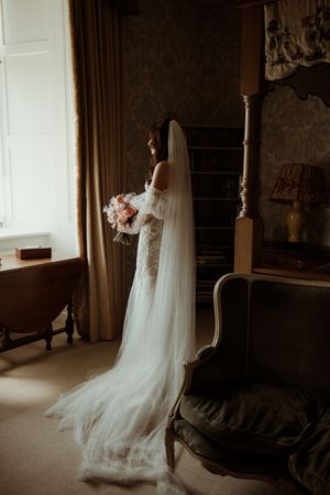 A bride in a long white lace gown and flowing veil stands by a window holding a pink bouquet in a dimly lit room.