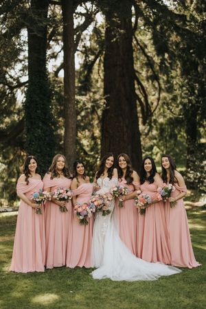 Bridal party in blush pink bridesmaid dresses pose together on lawn surrounded by tall trees in outdoor setting.