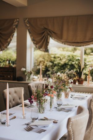 Elegant table setting with tufted chairs, white linens, delicate floral centerpieces and candles by sunlit windows.