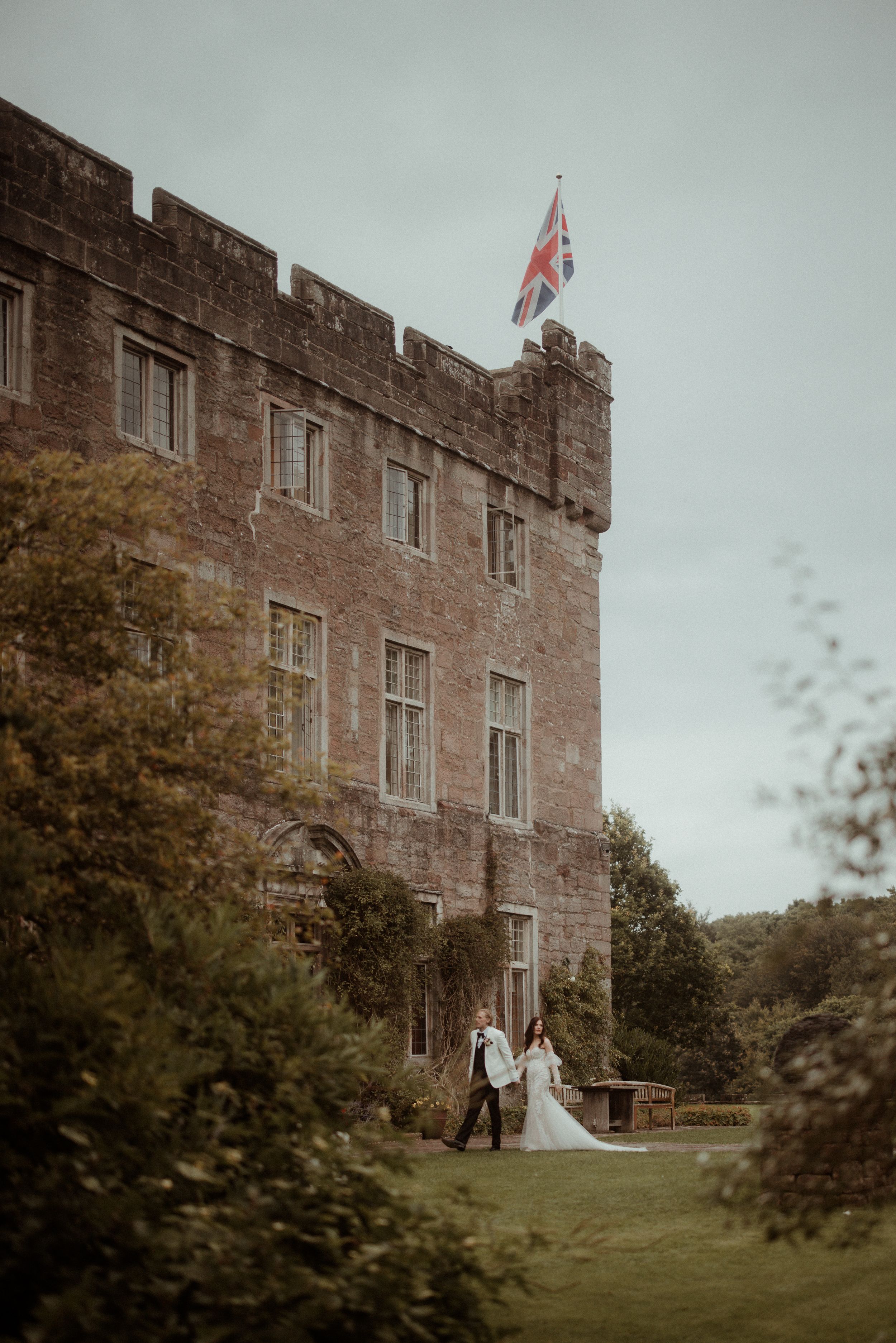 A couple stands outside a historic stone castle with a British flag flying above on an overcast autumn day.