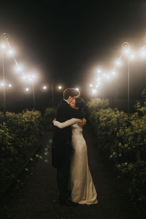 A couple embraces under romantic string lights at night in a garden setting with dark foliage surrounding them.