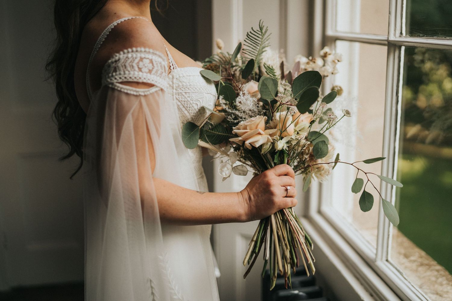 A bride in a white off-shoulder gown holds a rustic bouquet with greenery by a sunlit window. Taken by Grimsby photographers.