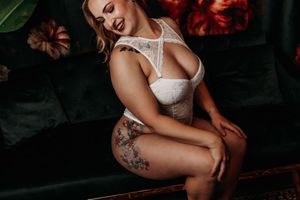 A model in white lace lingerie poses on a dark couch with moody red lighting and floral accents in the background. Boudoir Photo Studio MI