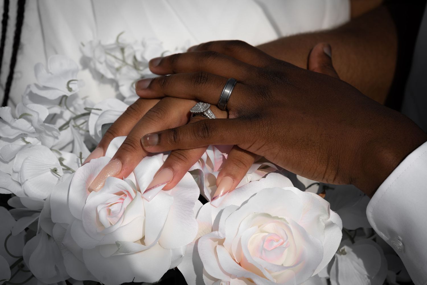 Hands wearing wedding rings rest on top of delicate pink roses and white fabric.