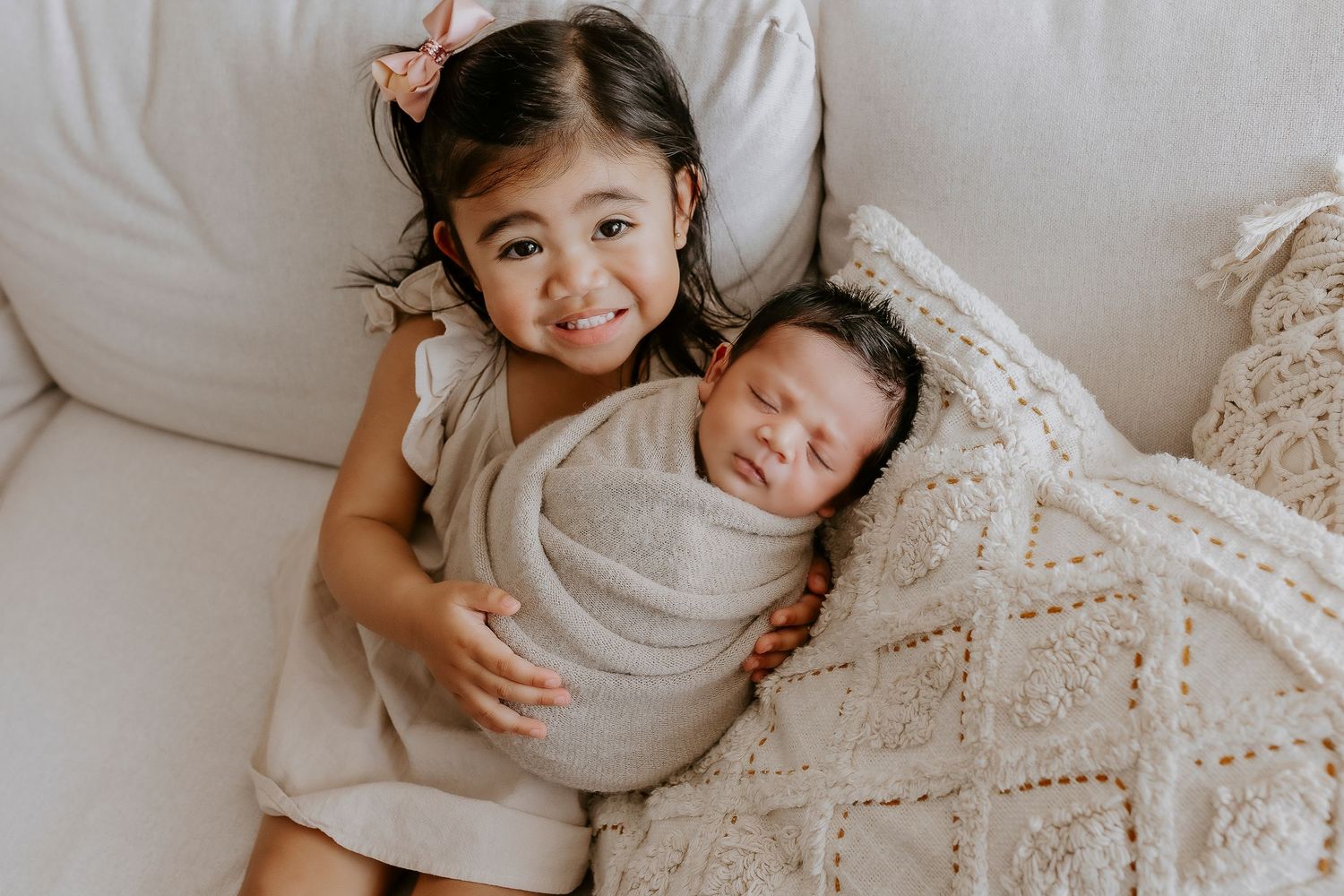 A tender moment as a sibling cuddles with a sleeping newborn on a couch with a patterned throw blanket.