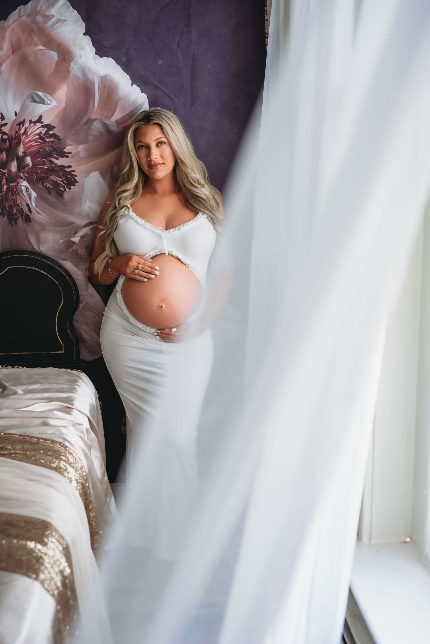 Maternity photo shoot featuring soft flowing white dress against romantic decor with delicate flower accents.