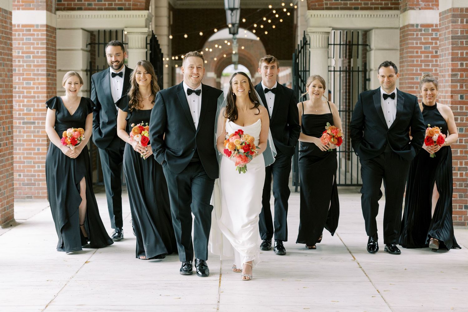 Wedding party in elegant navy blue dresses and black tuxedos poses together with bright coral bouquets.