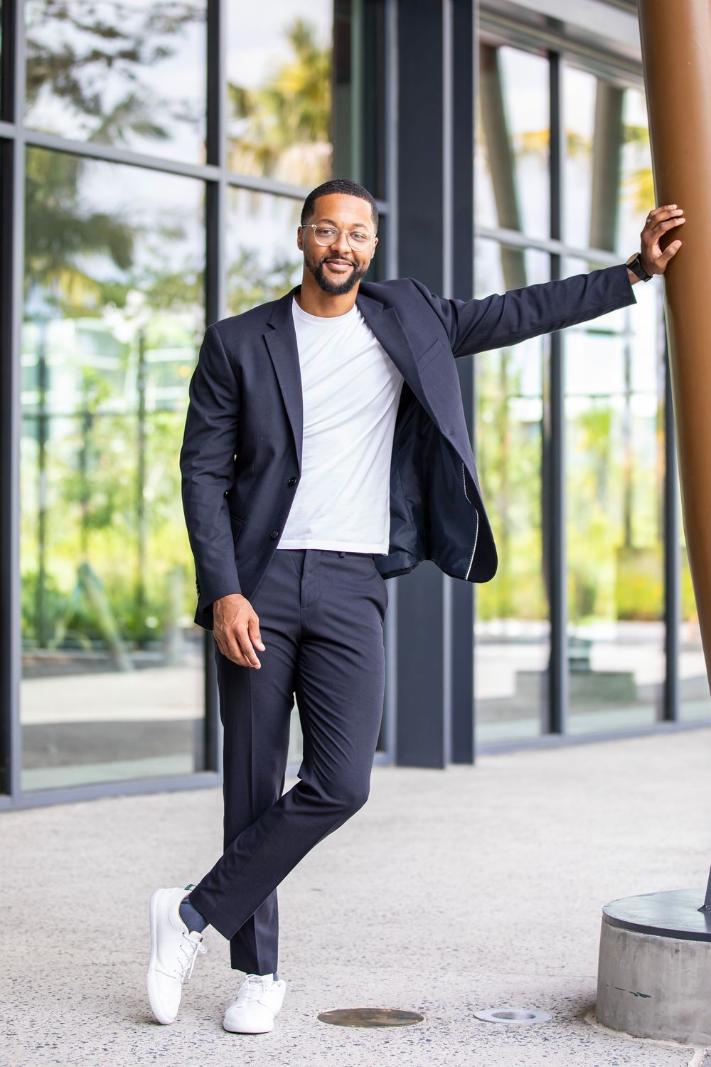 Person in navy suit and white sneakers poses playfully against modern building exterior.