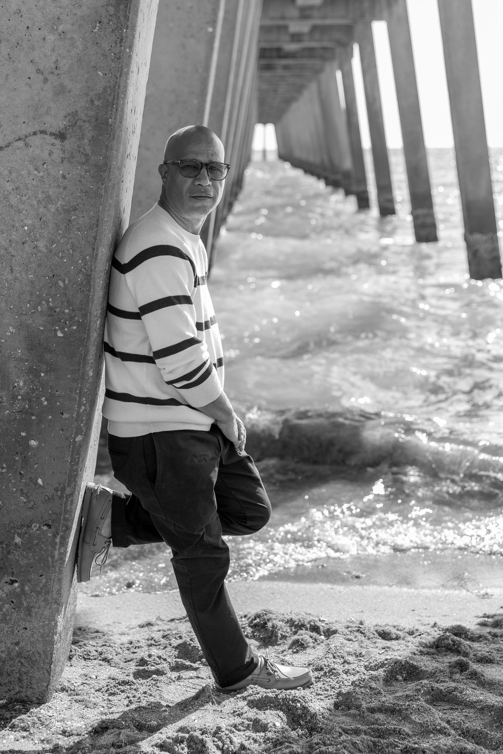 Black and white photo showing legs walking under a pier on sandy beach.