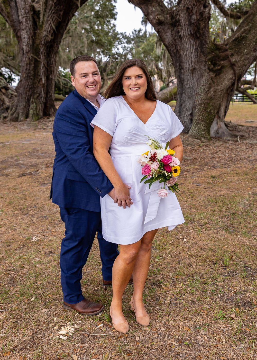 Couple in navy suit and white dress share romantic moment under oak trees.