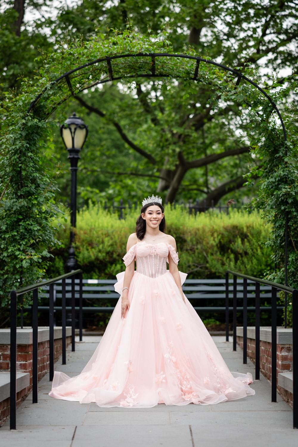 A flowing pink ball gown is showcased on a tree-lined path with garden benches.
