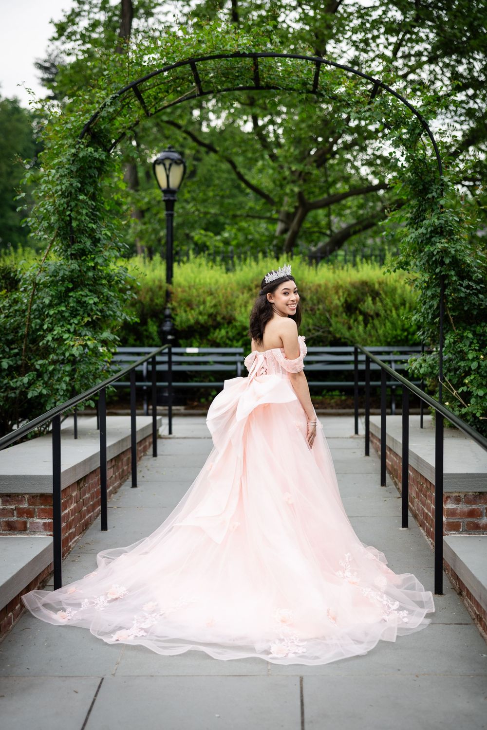 Pink ball gown dress spreads elegantly across a garden pathway with lamp posts.