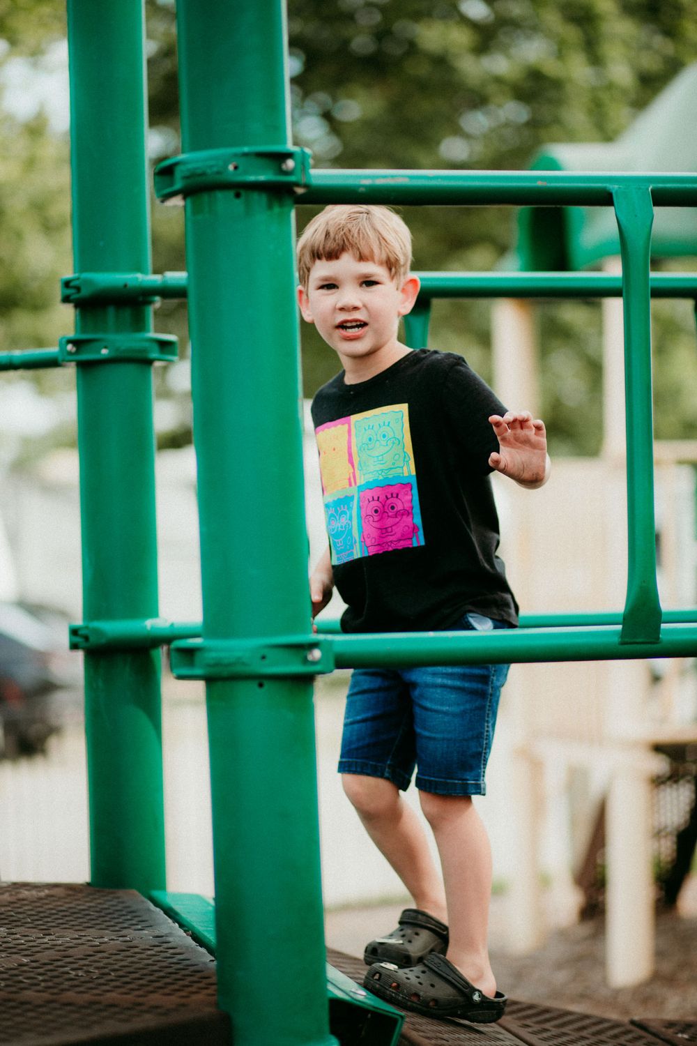 Child playing on green playground equipment wearing a black t-shirt and blue shorts on a sunny day.