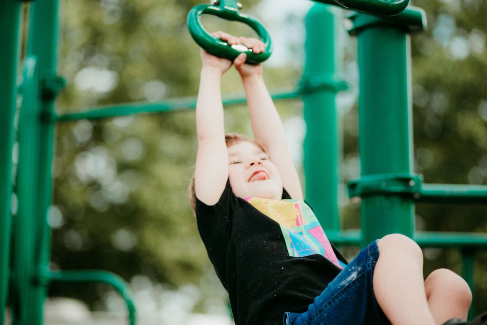 Small child in black shirt swings joyfully from green playground rings outdoors on a summer day.