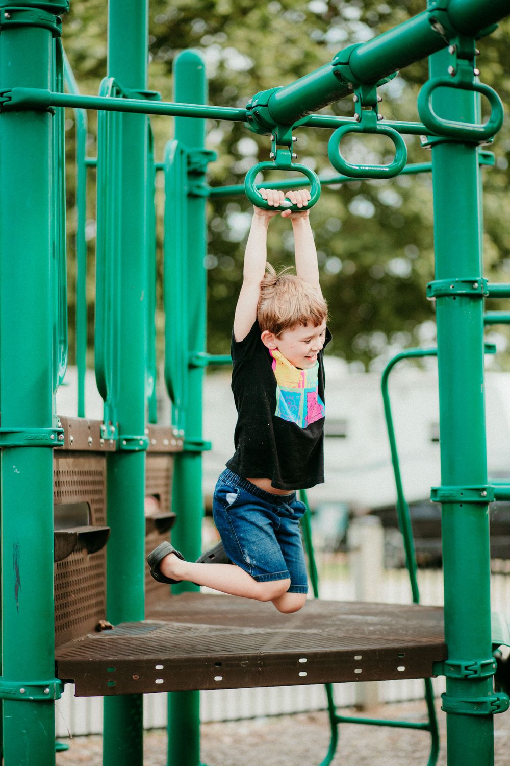 Young child in shorts and black shirt swings on green metal playground equipment bars at outdoor playground.