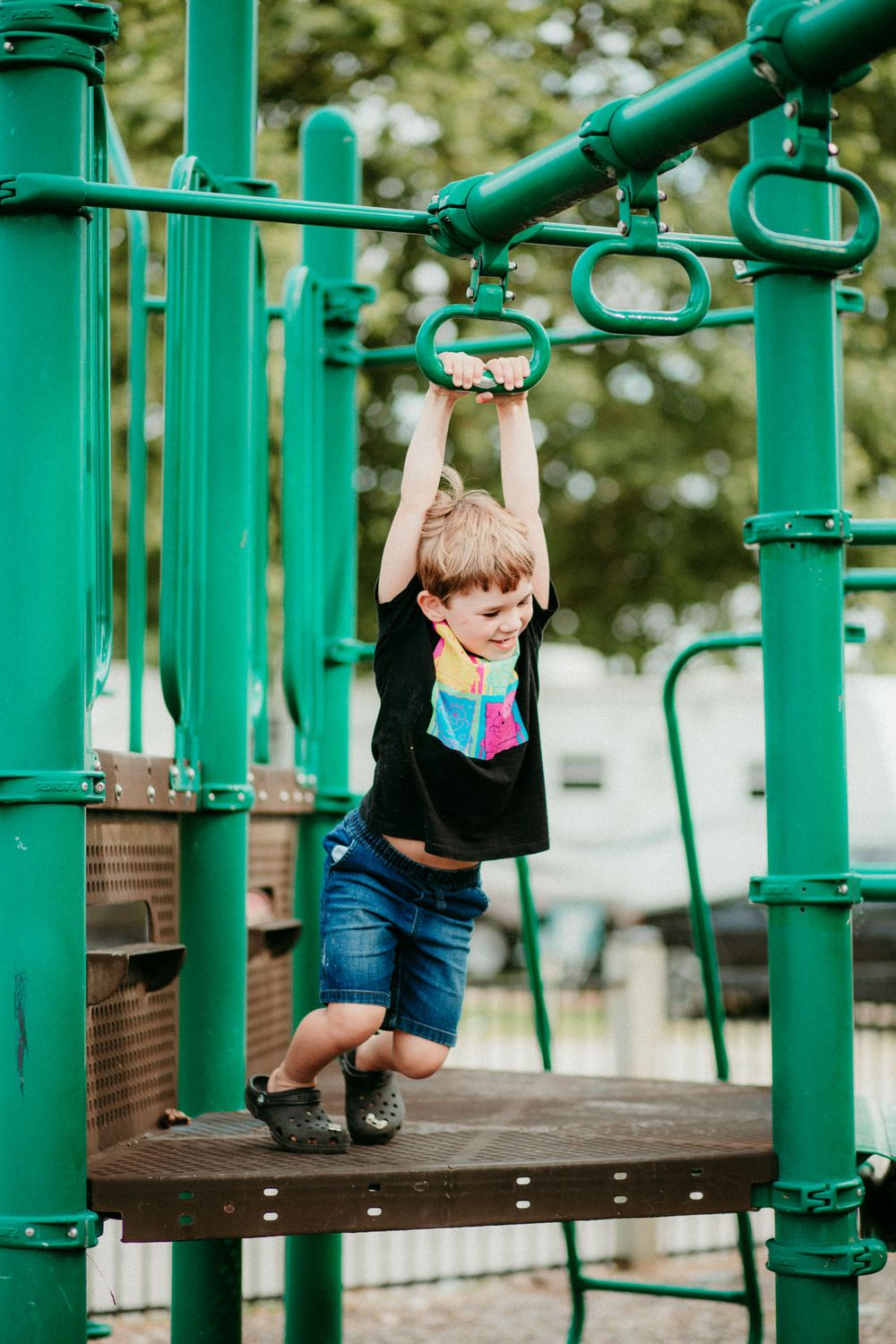 Child playing and hanging from bright green playground equipment on a sunny day at park.