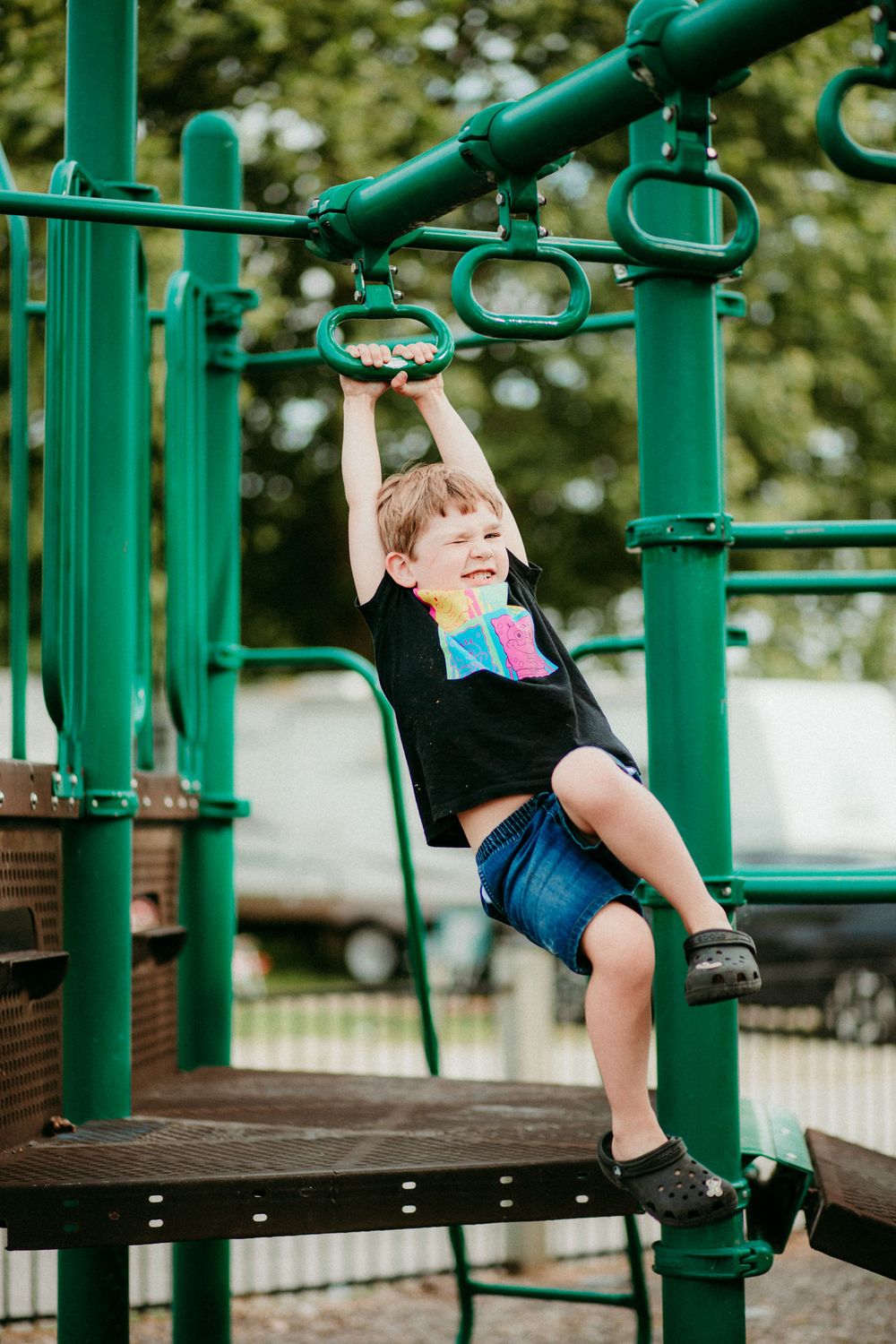 A child plays on green playground equipment, swinging and balancing across multiple metal bars at a park.
