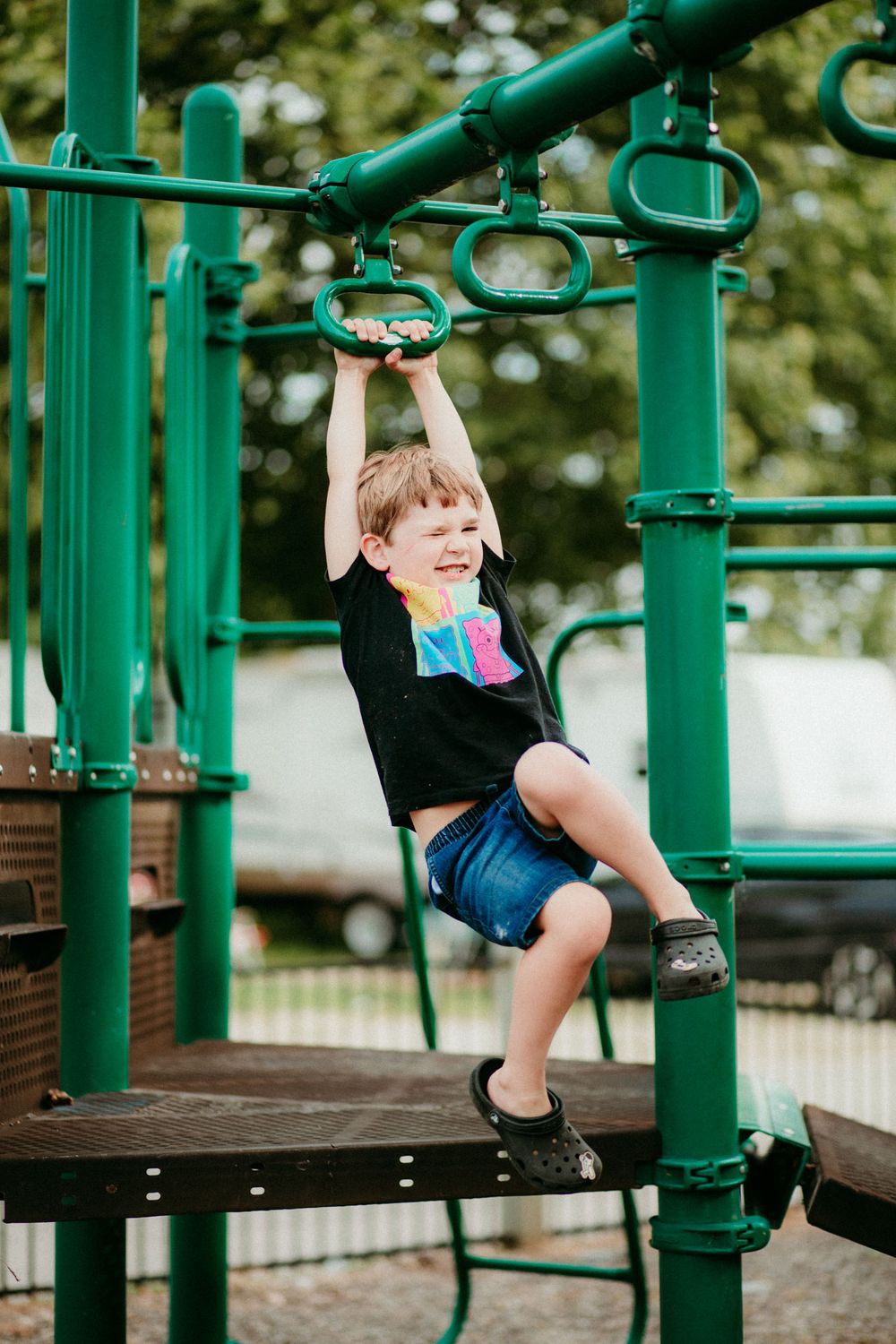 Young child playing and hanging from green playground equipment at a park on a sunny day.