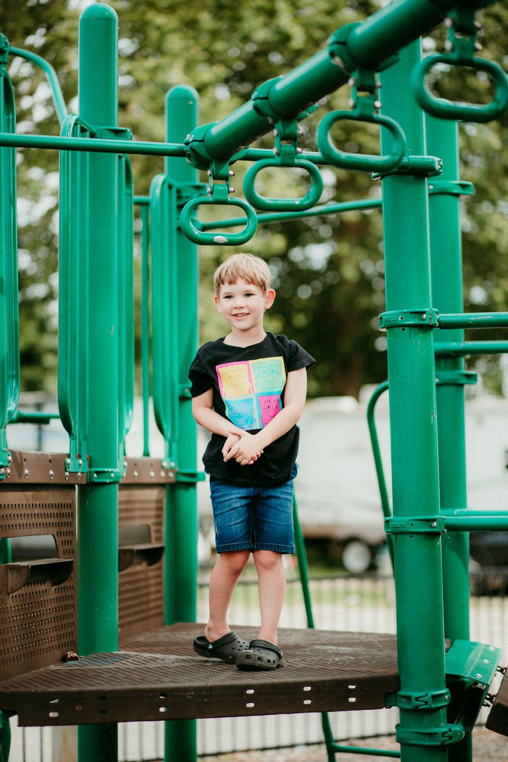 A young child stands on a bright green playground structure in jean shorts and sandals during a sunny day.