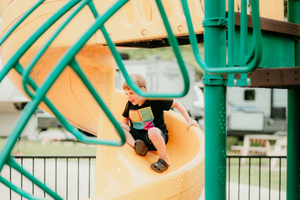 Child climbs up a yellow and turquoise spiral slide at a playground on a sunny day.