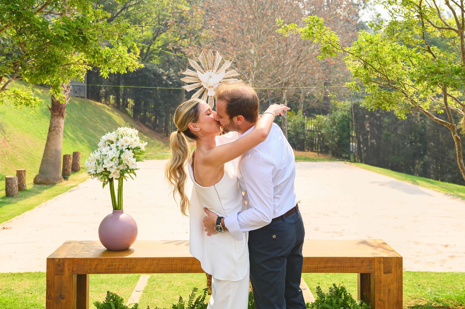 A romantic wedding couple embraces and kisses on a wooden bench in a sunlit garden setting with a floral centerpiece.