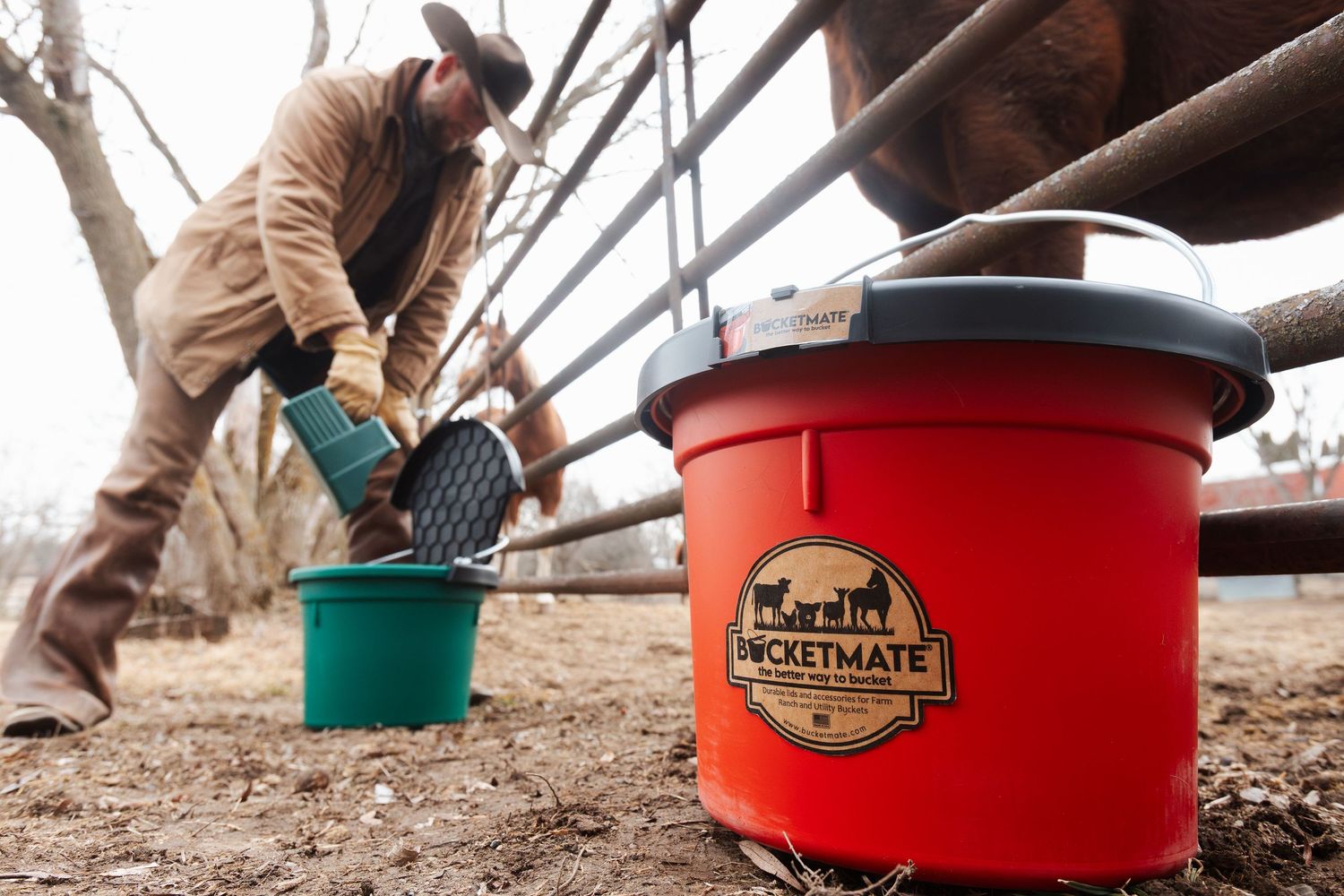 A red Bucketmate container sits in the foreground while someone works with cattle in a ranch setting.