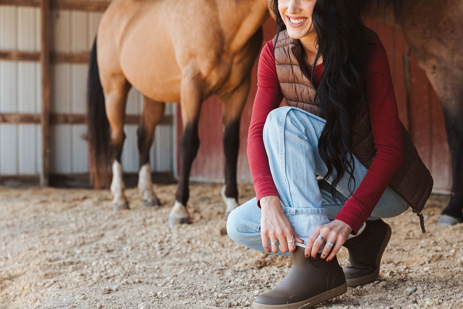 Person in burgundy sweater and jeans kneels to lace up boots in a horse stable with a brown horse in background.