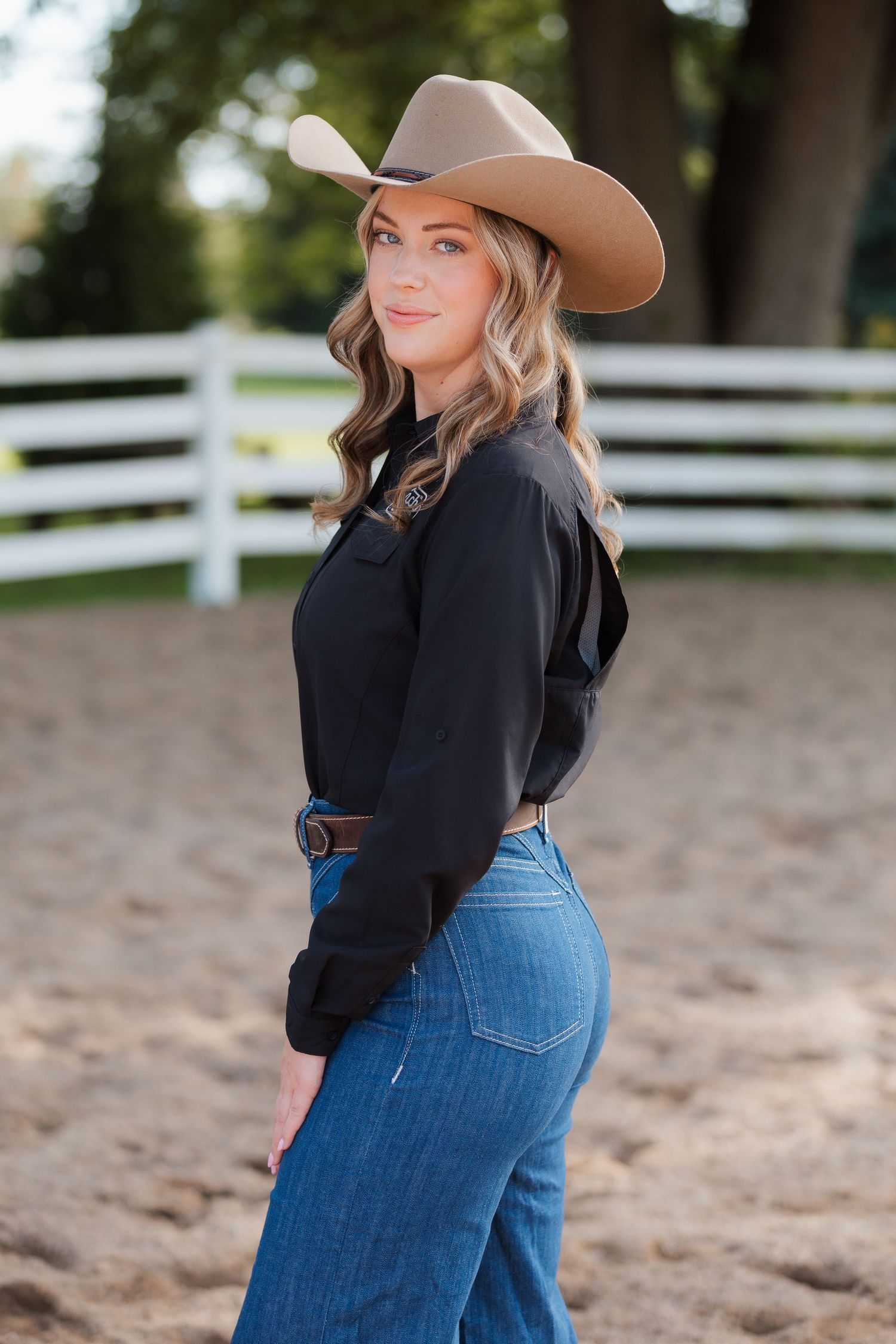 Young woman in cowboy hat, black shirt, and jeans poses at a ranch with white fence backdrop.
