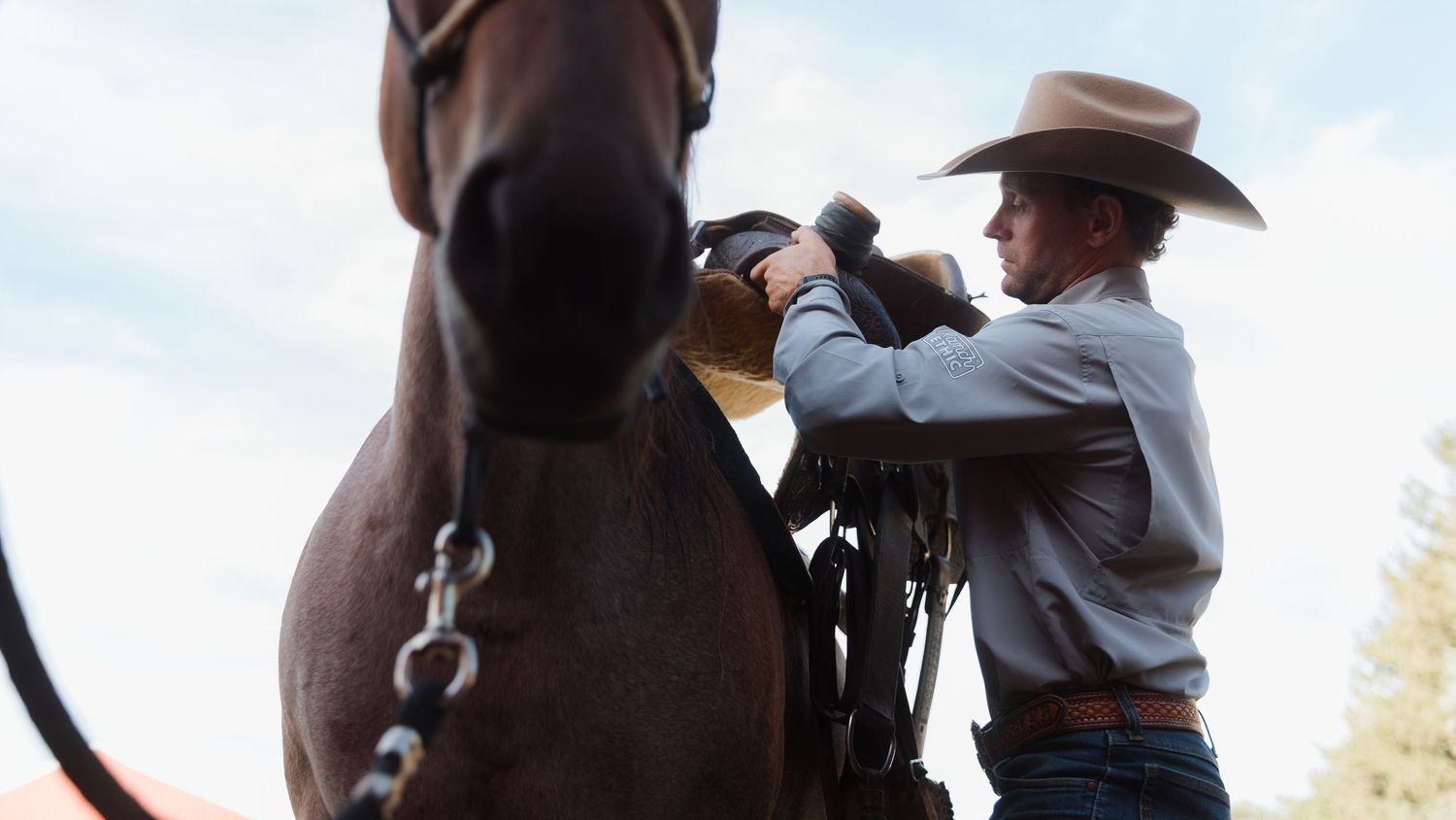 A cowboy in a tan hat adjusts the bridle on a dark brown horse outdoors in a rustic ranch setting.