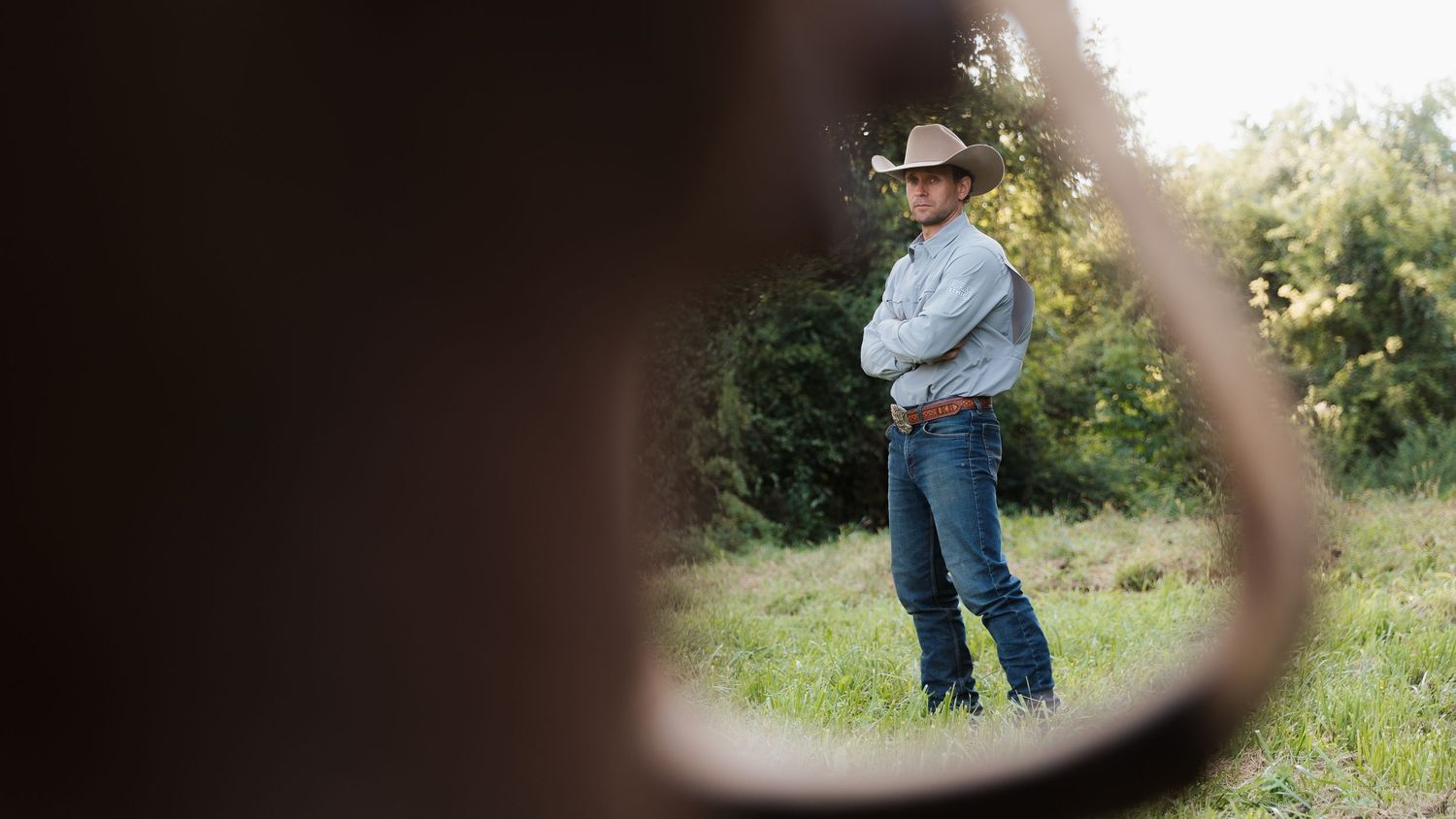 A cowboy in a hat and jeans stands with arms crossed in a green field, viewed through a circular frame.