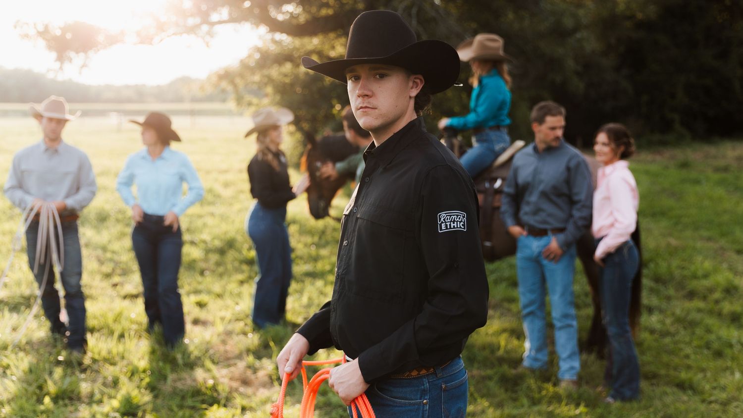 Young cowboy in black hat and jacket stands in foreground with group of ranchers and horse in sunny field.