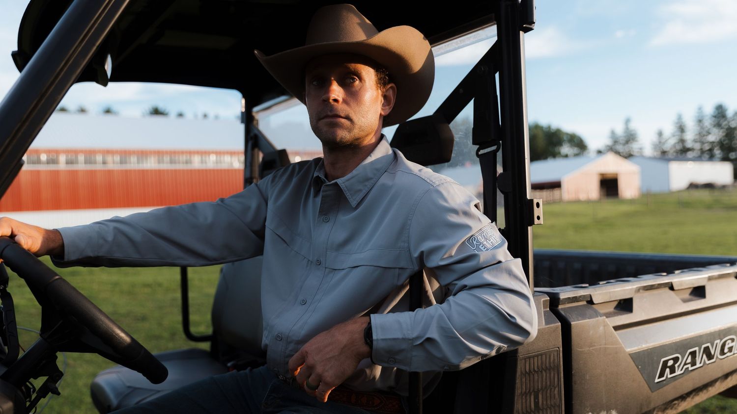 A young man in a cowboy hat sits in a ranch utility vehicle on a green farm with red barn in background.