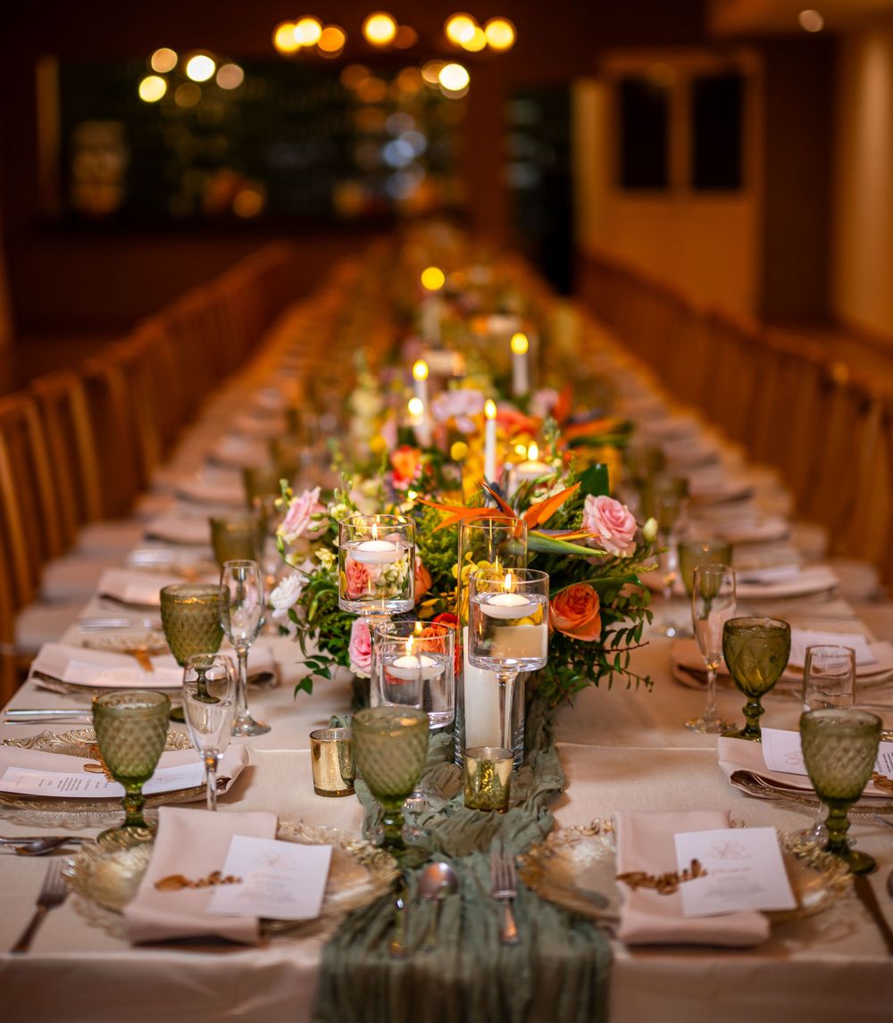 Long banquet table set for wedding reception with floral centerpieces, candles and place settings in warm lighting.