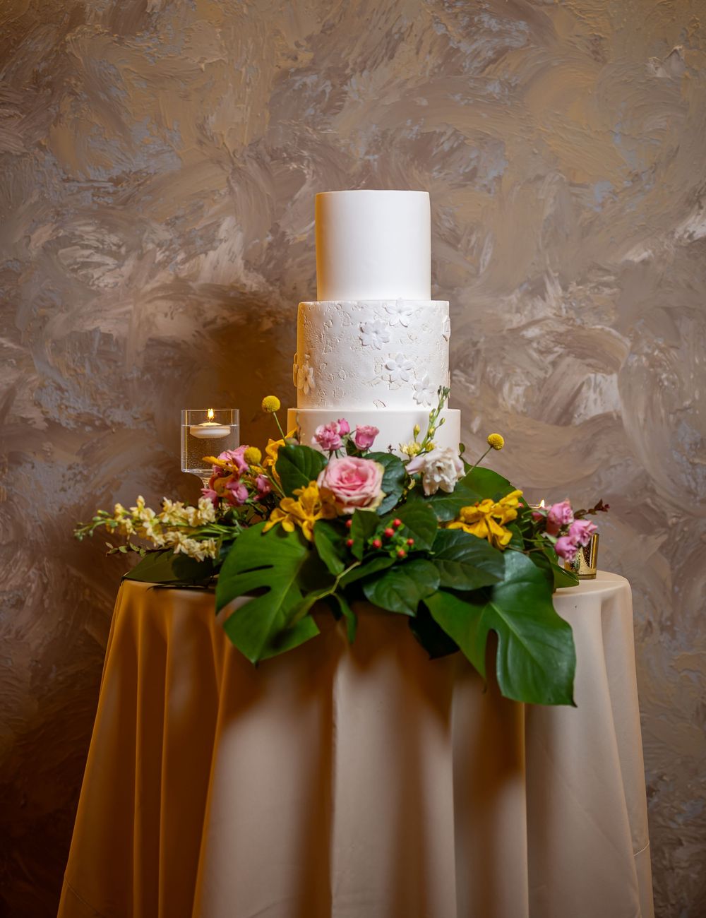 White two-tier wedding cake decorated with pink roses and tropical leaves on a champagne-colored tablecloth.