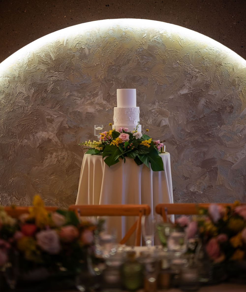 Elegant white wedding cake displayed on draped table with floral decorations against dramatic illuminated wall.
